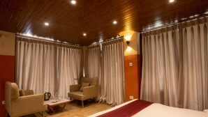 Habitación de hotel con techo de madera, dos sillones, una mesita con hervidor y vasos, una cama con manta roja y cortinas en las ventanas.