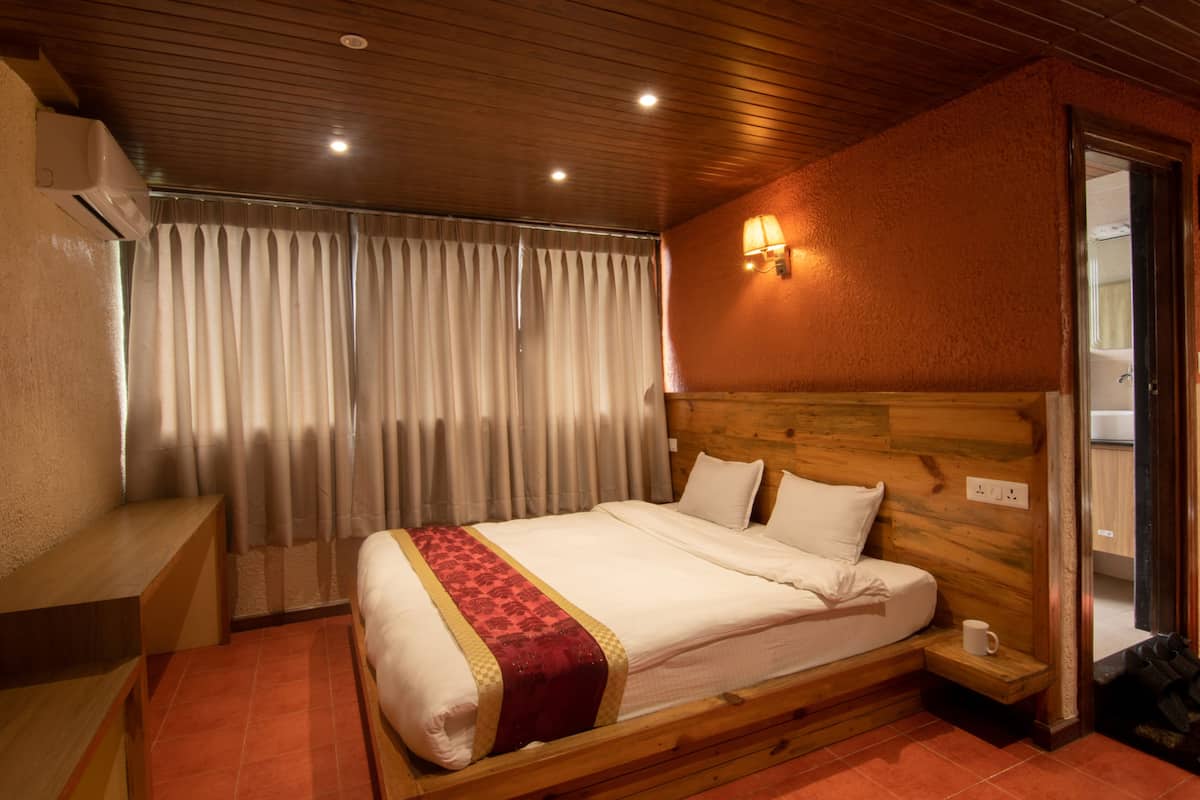 Una cama con estructura de madera, ropa de cama blanca y un detalle rojo, una mesita de noche con una taza, y un banco de madera.