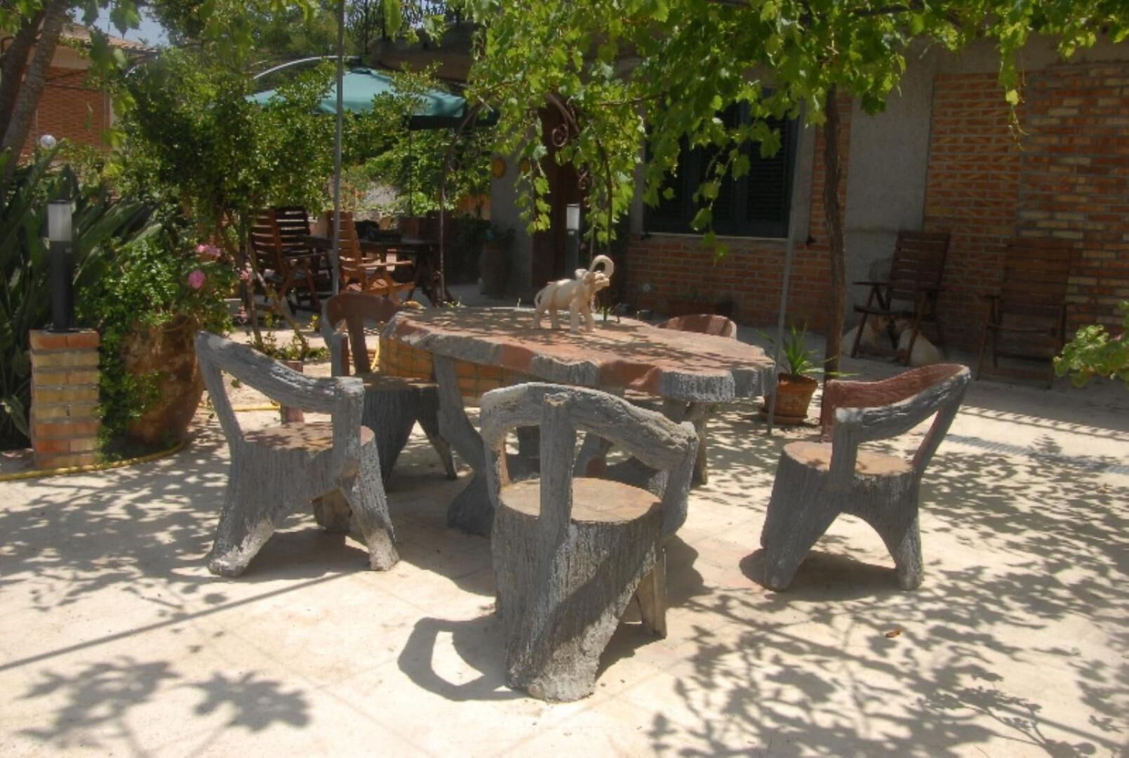 An outdoor patio with a round stone table and six rustic chairs, surrounded by potted plants and a brick wall.