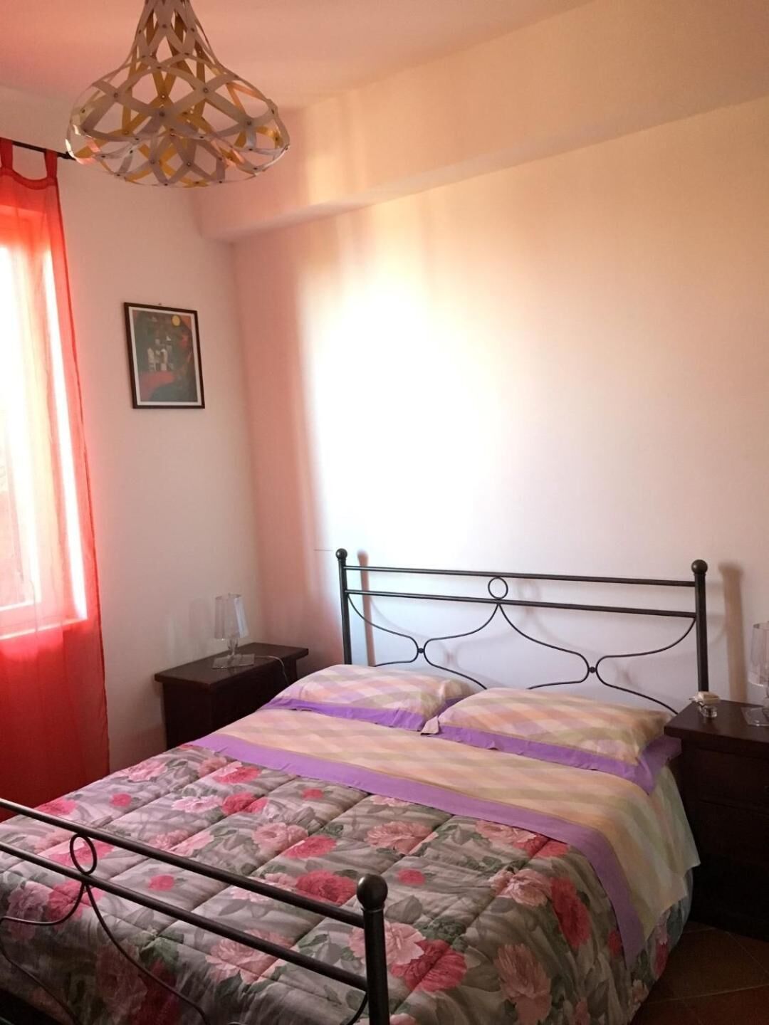Un dormitorio con cama, mesita de noche, lámpara, un cuadro en la pared y una ventana con cortinas.