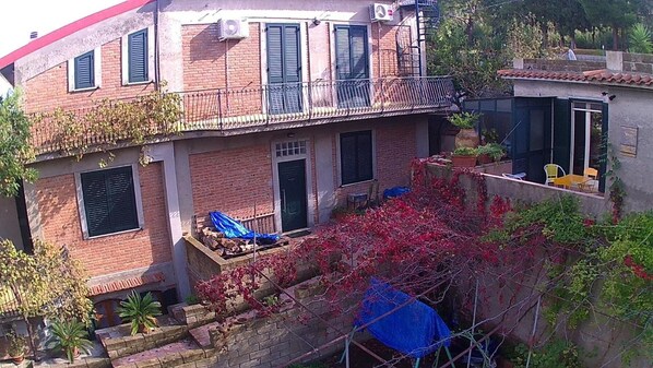A two-story building with a balcony, a door, and a small outdoor area with plants and a blue tarp.