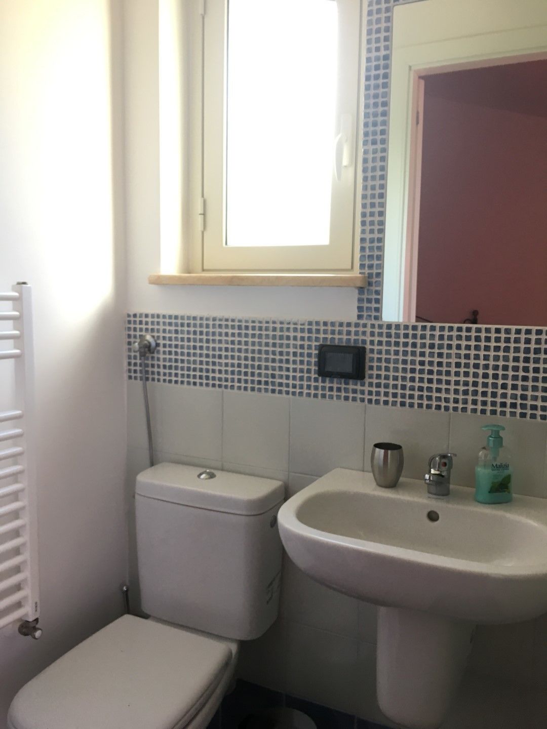 Un baño pequeño con inodoro, lavamanos y un ventanal sobre el lavamanos.