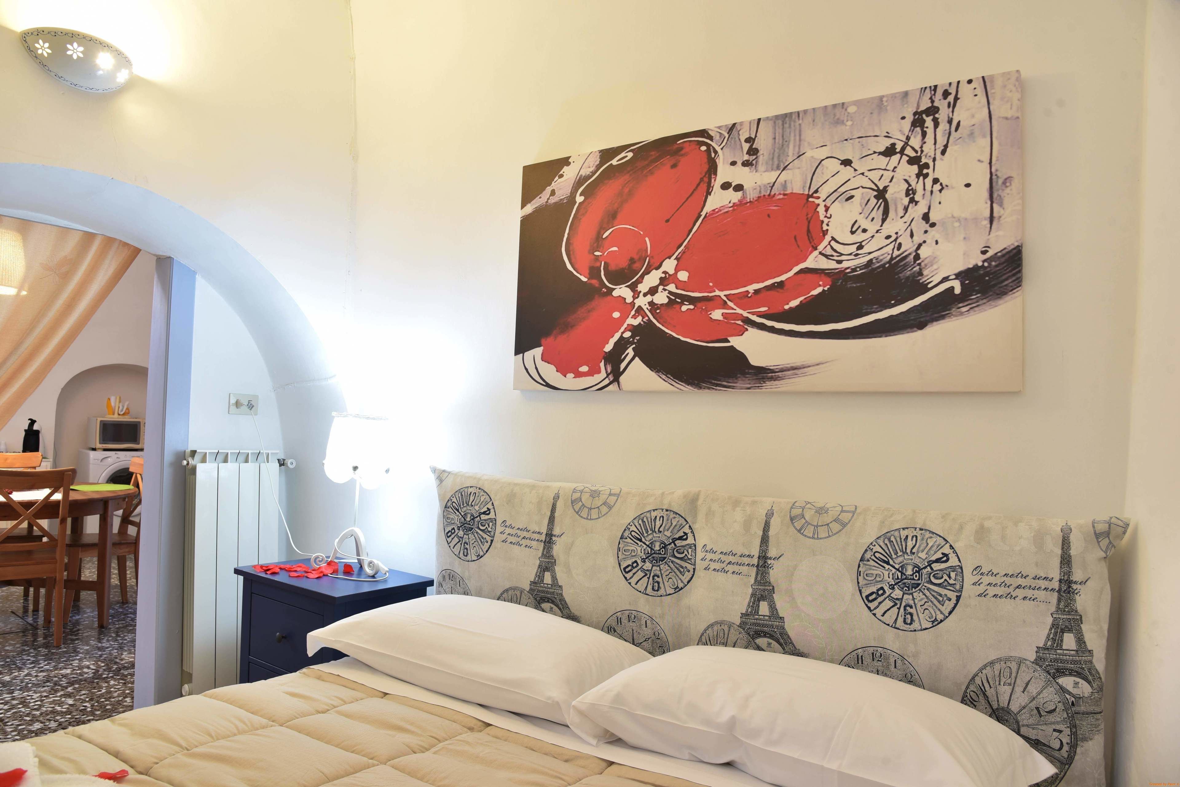 A hotel room with a bed, a nightstand, a desk with a chair, and a painting of the Eiffel Tower and clocks on the wall.