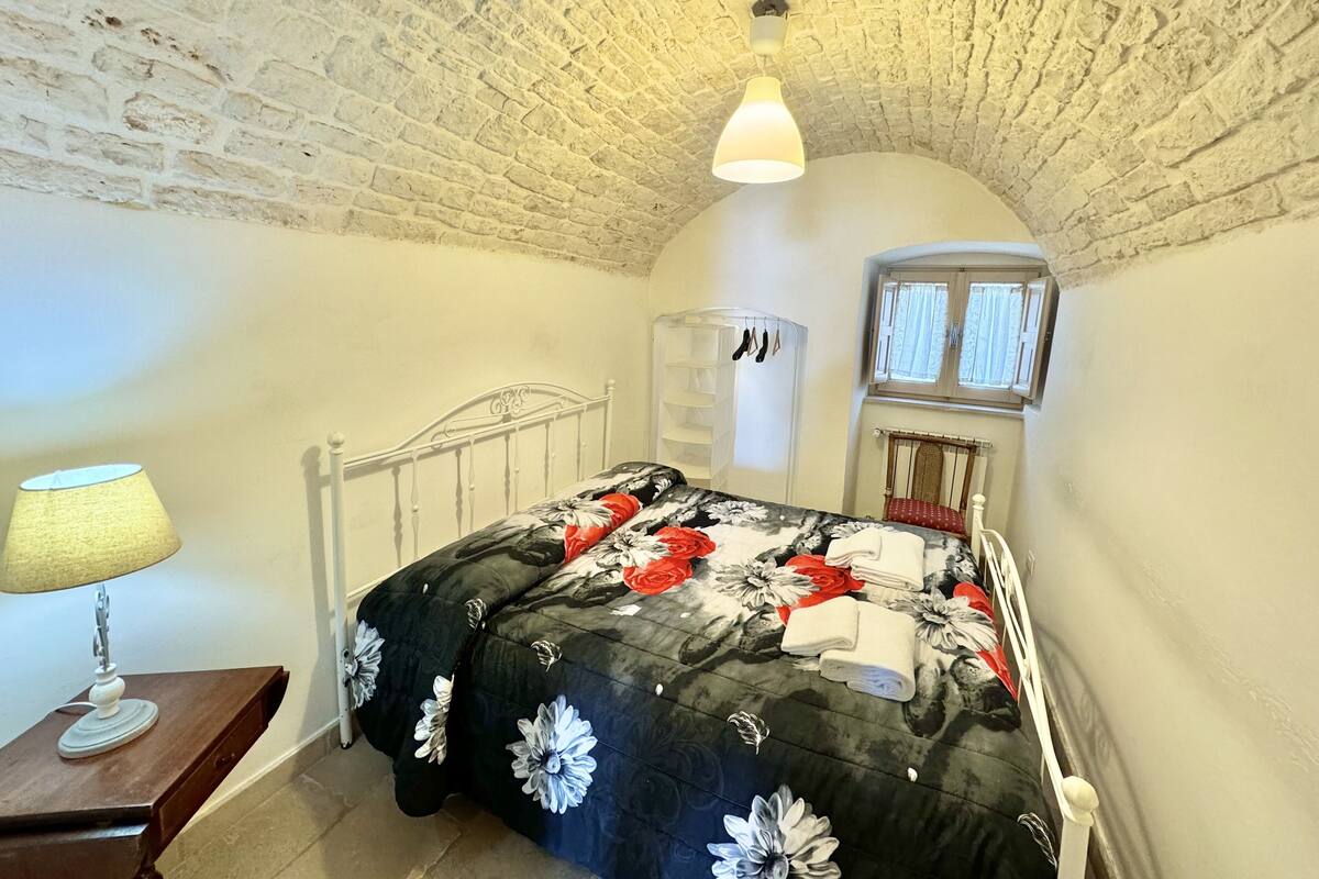 A bedroom with a white brick ceiling, a bed with a floral duvet, a bedside table with a lamp, and a window with curtains.