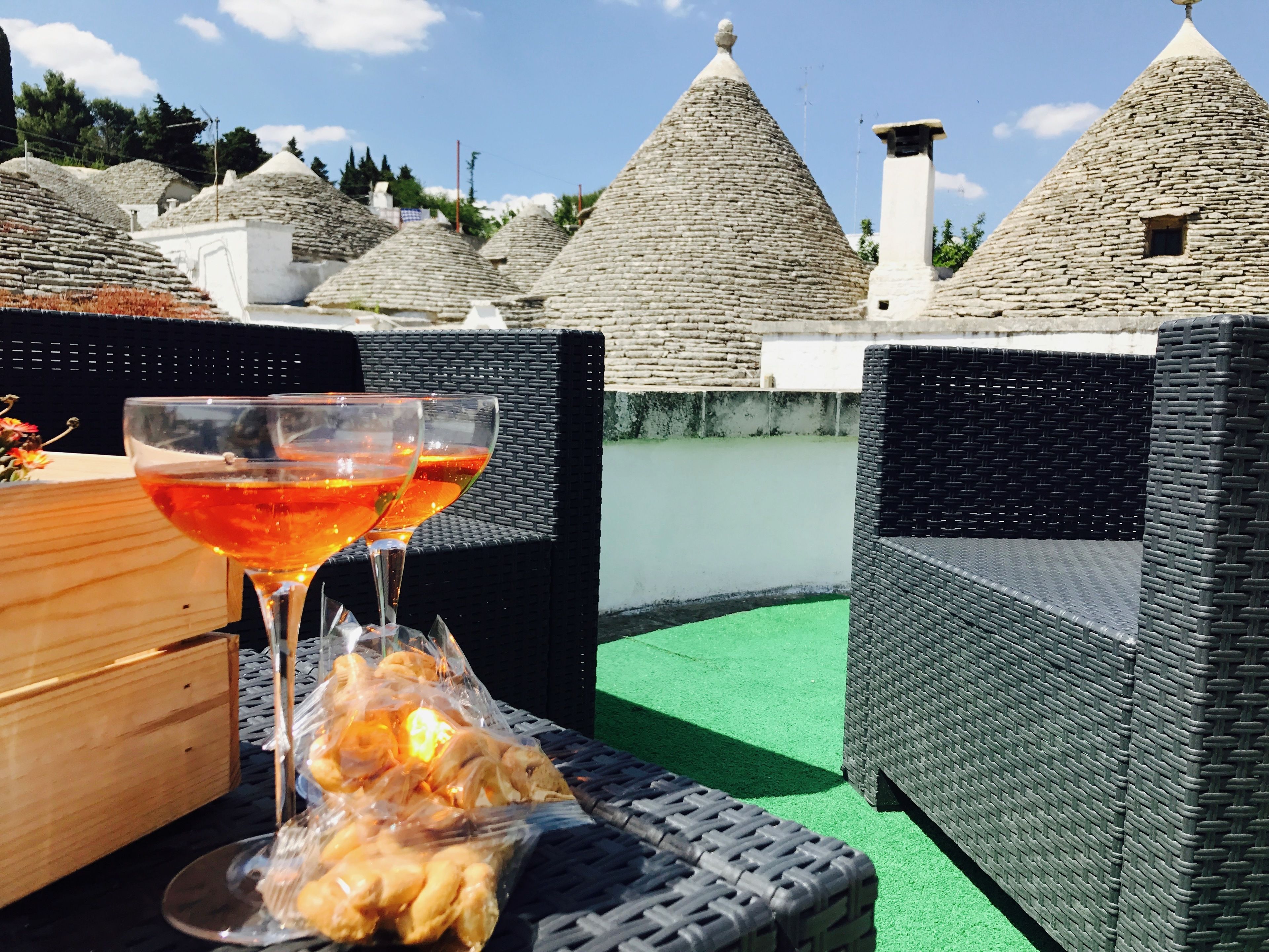Una mesa de mimbre con una copa de vino tinto y una bolsa de plástico con pasteles, frente a edificios de piedra con tejados cónicos.