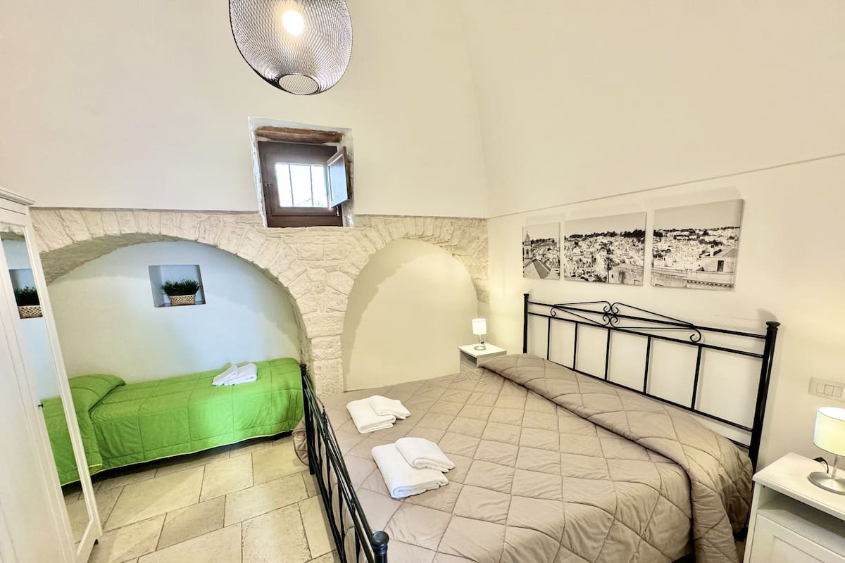 A bedroom with a bed, a green sofa, a stone archway, a nightstand, and a wall with three black-and-white photographs.
