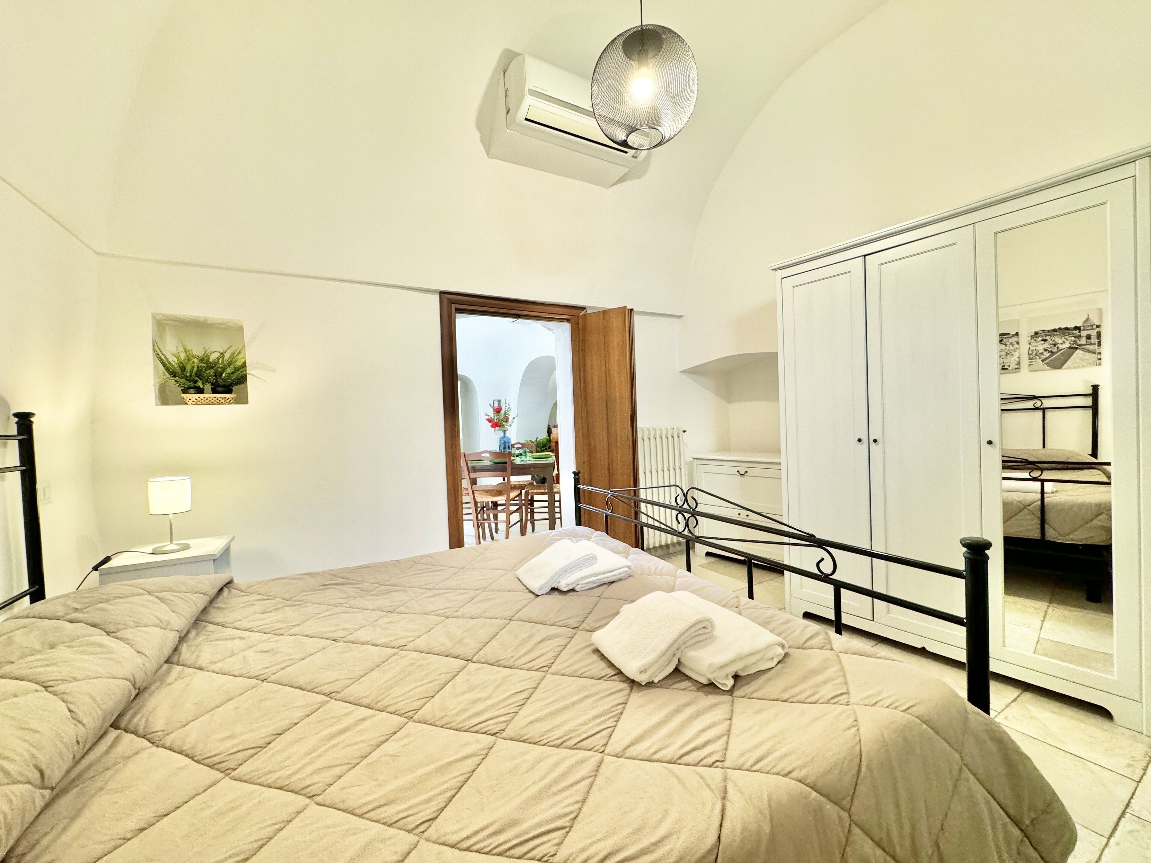 Quarto com uma cama grande, uma mesa de cabeceira com luminária, guarda-roupa e vista para uma varanda com área de jantar.