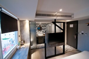 Habitación de hotel moderna con cama, televisión, baño pequeño con lavamanos y ventana con vista a la ciudad.