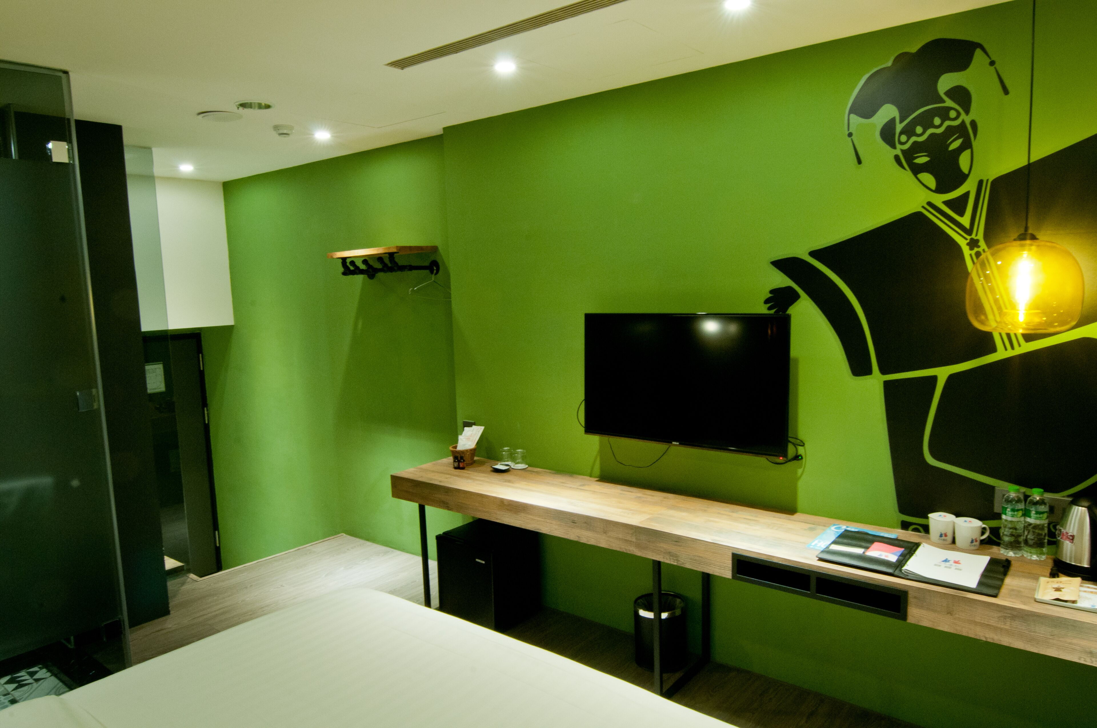Une chambre d’hôtel avec un mur vert, un téléviseur à écran plat fixé au mur, un bureau en bois avec un ordinateur, une poubelle et une petite étagère avec une tasse et une bouteille.
