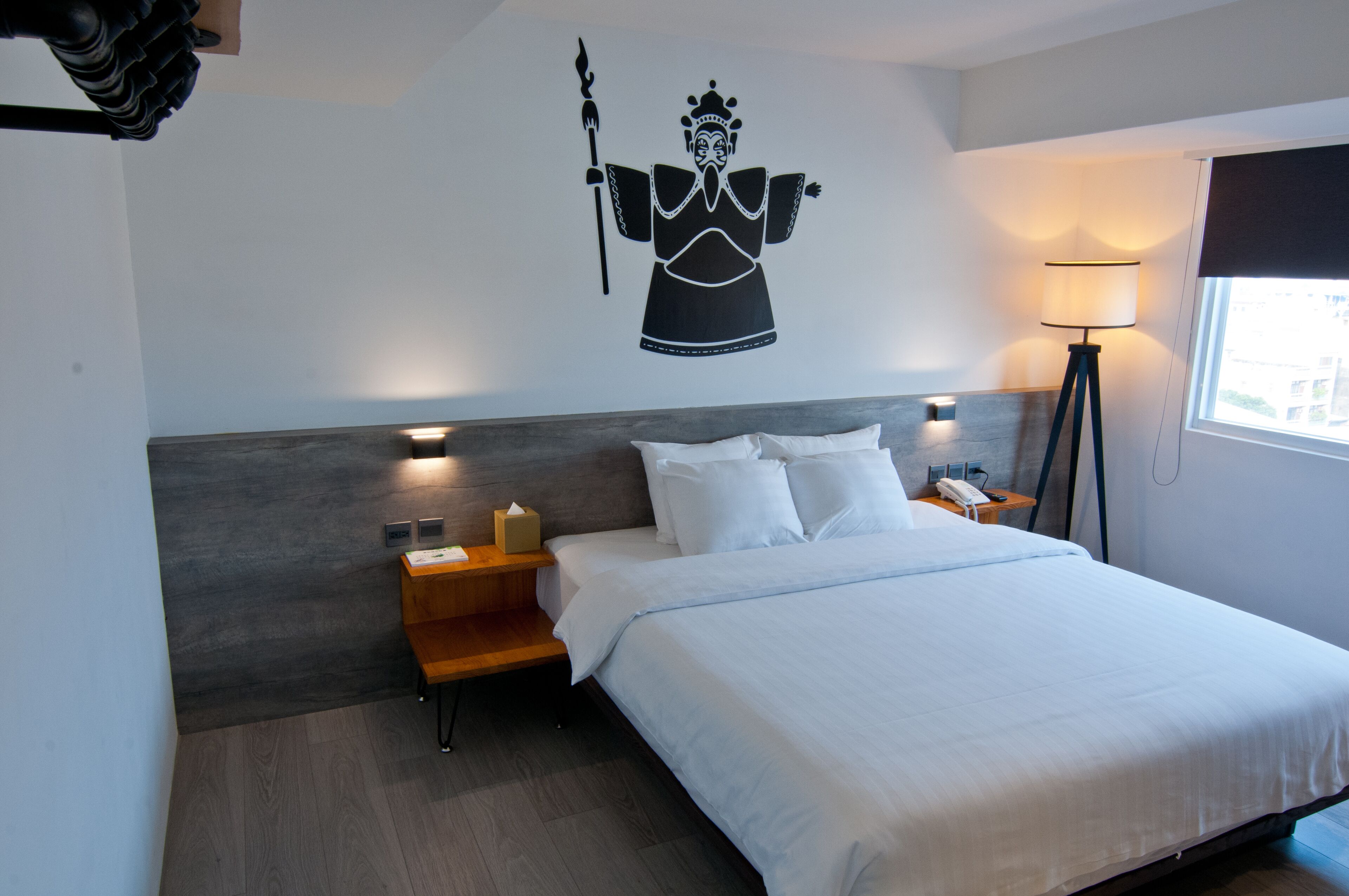A hotel room with a large bed, a nightstand with a lamp, a wall-mounted artwork, and a window with a view.