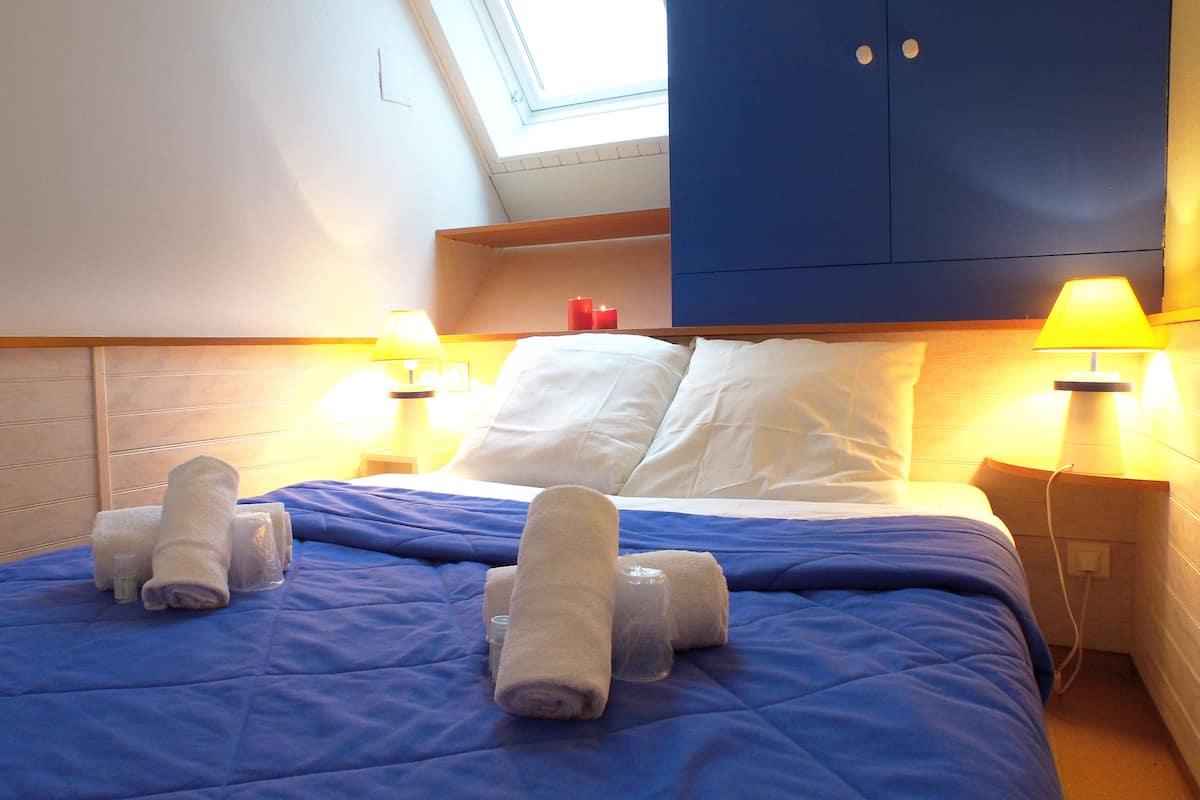 Et hotelværelse med en seng, to sengeborde, to læselamper, et blåt sengetæppe og et ovenlys over sengen.