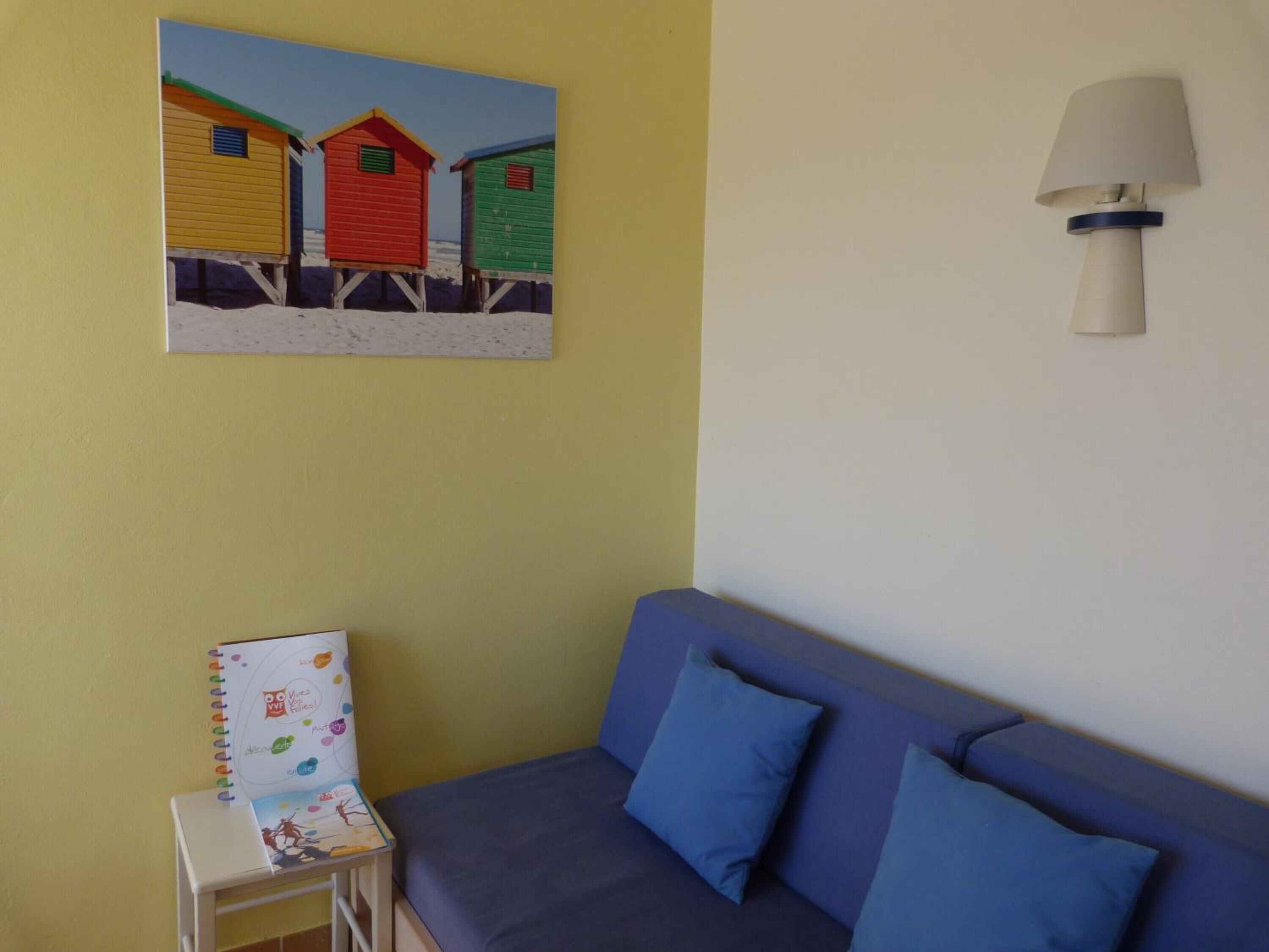 Une pièce avec un mur jaune, un canapé bleu assorti de coussins, une petite table blanche avec un carnet, et un tableau représentant des cabanes de plage aux couleurs vives.