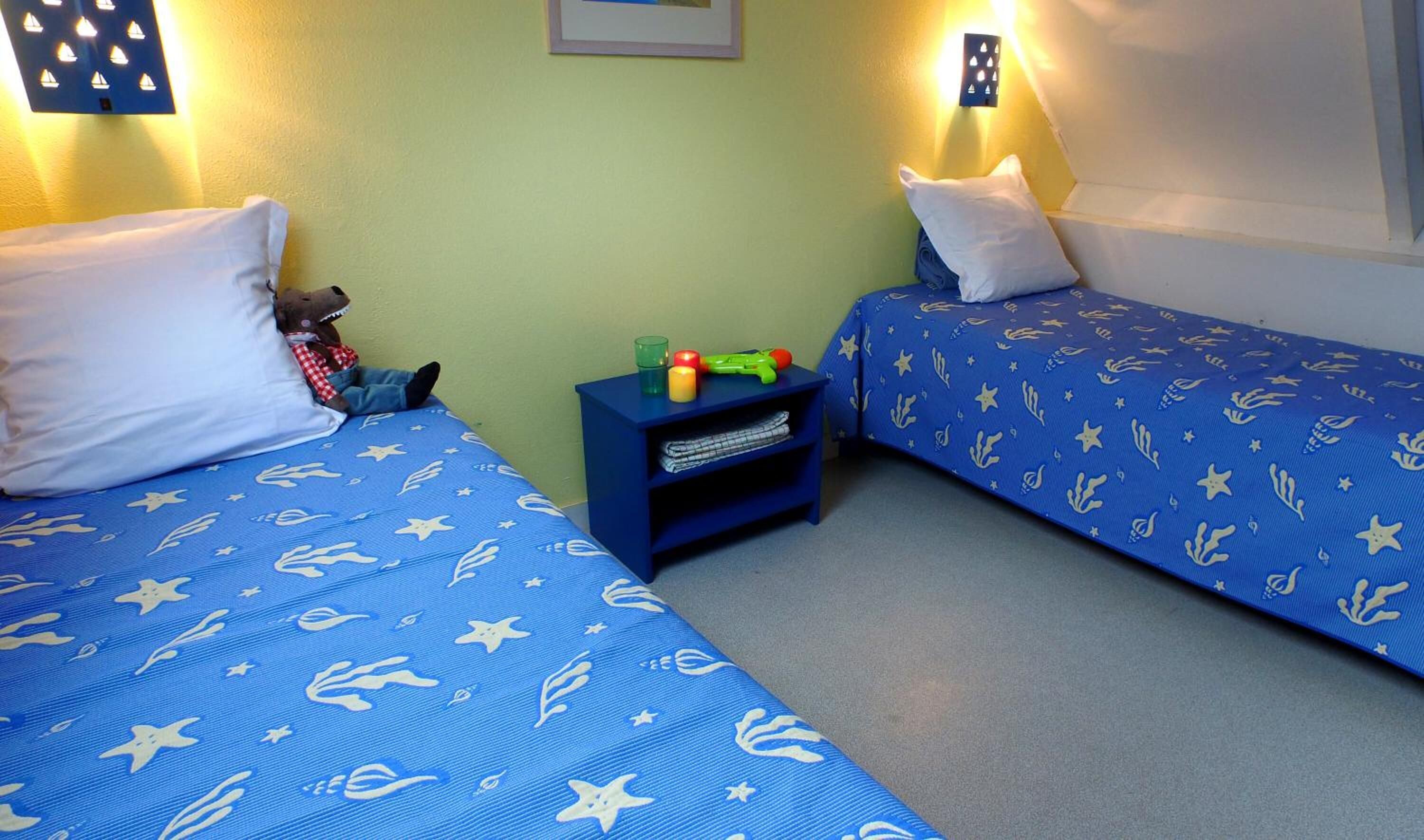 A room with two beds, each with a blue bedspread featuring white starfish and seaweed patterns. There is a small blue nightstand between the beds with a green cup, a red candle, and a toy.