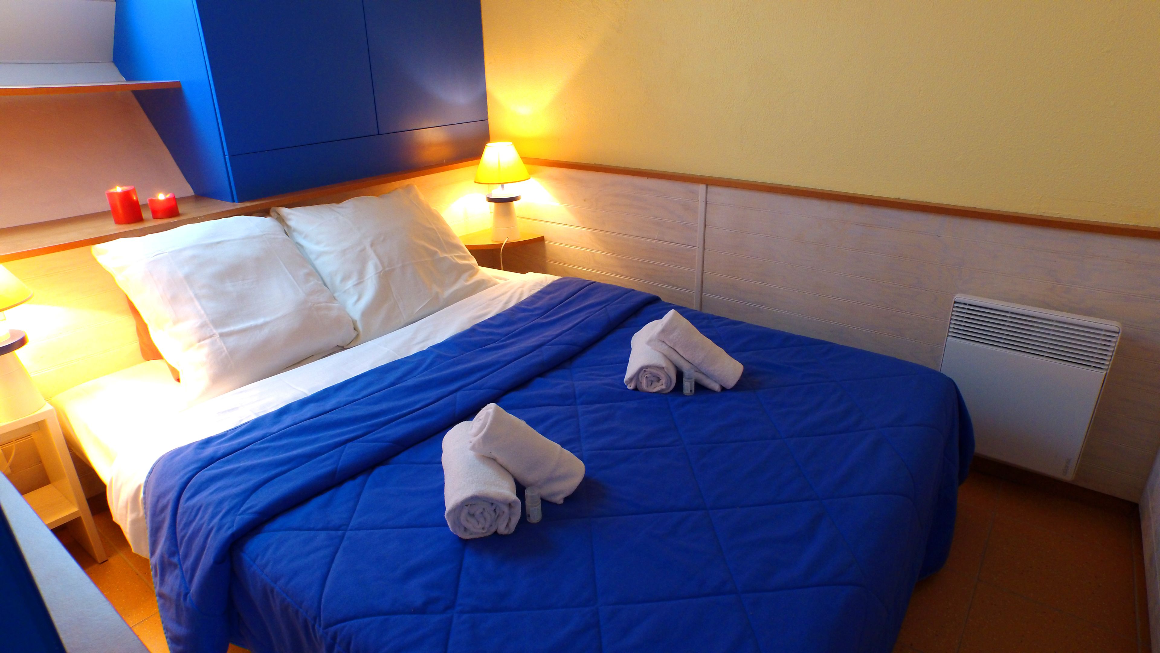 A neatly made bed with a blue quilt, white pillows, and two rolled towels. There are two bedside lamps with red candles, and a white vent on the wall.