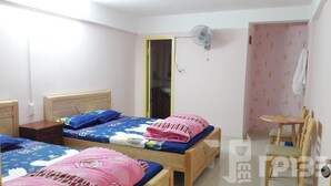 Une chambre à coucher avec deux lits en bois, une petite table, une chaise, un ventilateur et une porte menant à la salle de bain.