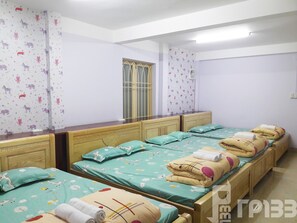 A hotel room with four single beds, each with a teal bedspread and matching pillows. The room has white walls, a wooden headboard, and a window with a metal grill.