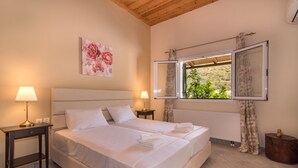 Uma cama de casal com lençóis brancos, duas mesas de cabeceira com abajures, um quadro floral na parede e uma janela com cortinas.