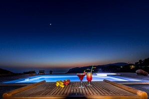 A wooden deck with a pool, two drinks, and fruit.