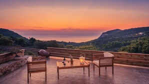 Terraço com mobiliário de madeira, mesa com uma garrafa e uma abacaxi, e vista para o mar ao pôr do sol.