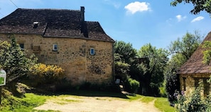 A stone building with a dirt path leading to it, surrounded by greenery and trees. - Sur le Chemin des Sens (Azerat)