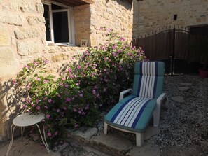 A stone building with a window, a chair with striped cushions, and pink flowering plants. - Sur le Chemin des Sens (Azerat)
