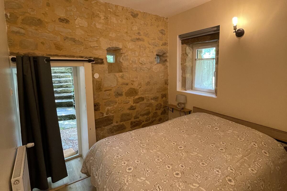 A bedroom with a stone wall, a bed with a floral bedspread, a window with a view, and a wall-mounted light fixture.