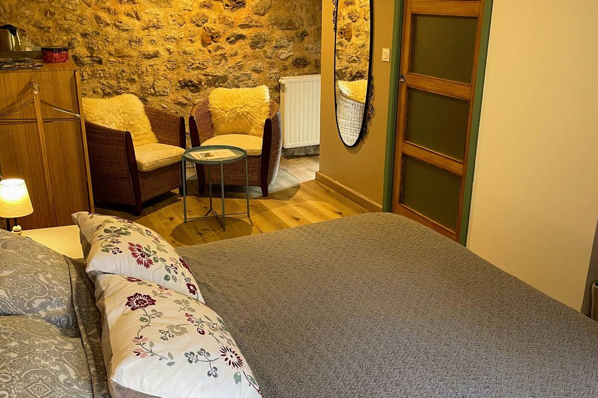 A bedroom with a large bed, a chair, a small table, a mirror, and a stone wall.