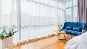 A room with a large window covered by sheer curtains, a blue sofa, a small wooden table with a vase of flowers, and a potted plant on the windowsill. - Mishe TEN Boutique Inn (Shanghai)