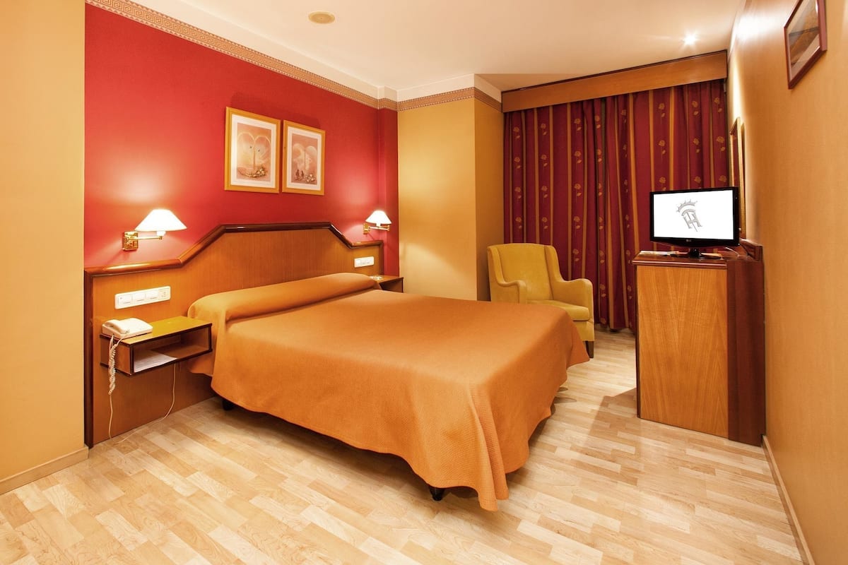 A hotel room with a bed, bedside lamps, a desk with a telephone, a chair, a television, and a curtain.