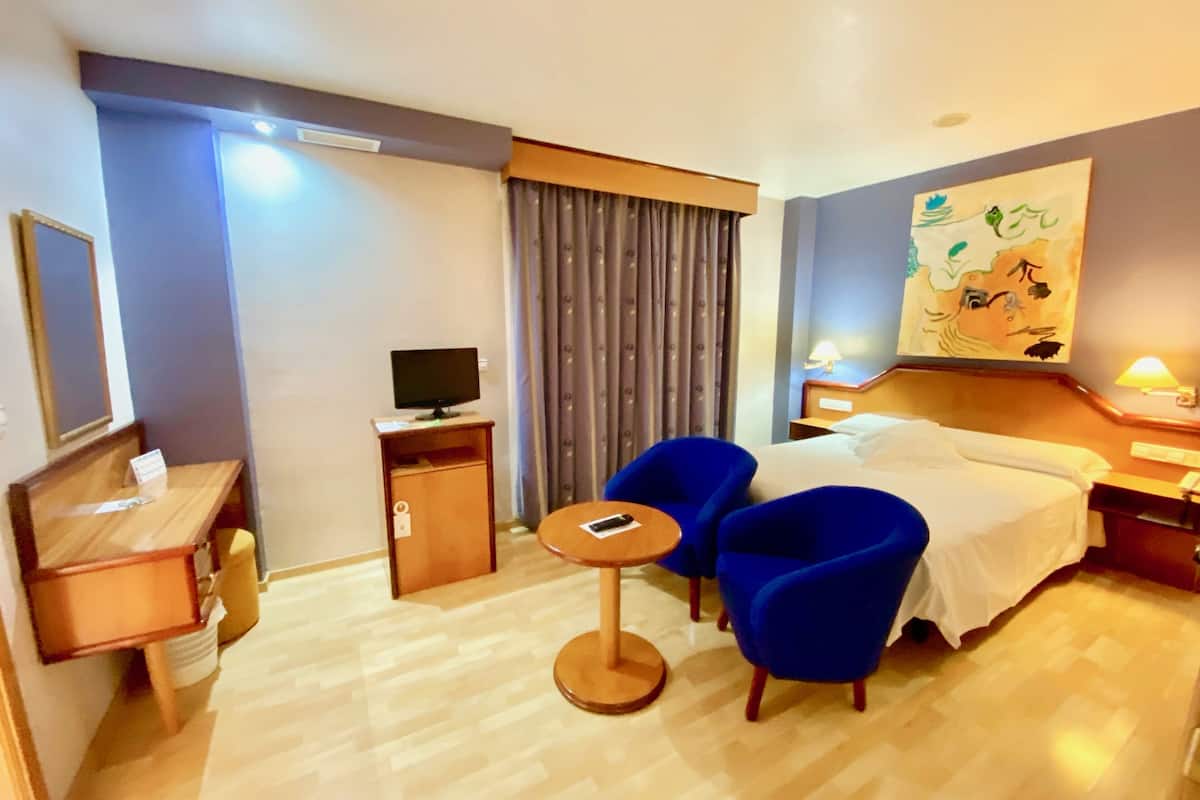 A hotel room with a bed, a desk, a chair, a TV, and a painting on the wall.
