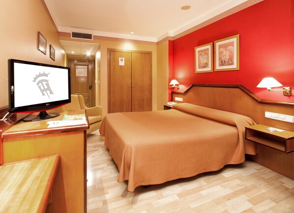 Habitación de hotel con cama, escritorio con monitor de computadora, silla, mesita de noche y dos lámparas de pared.