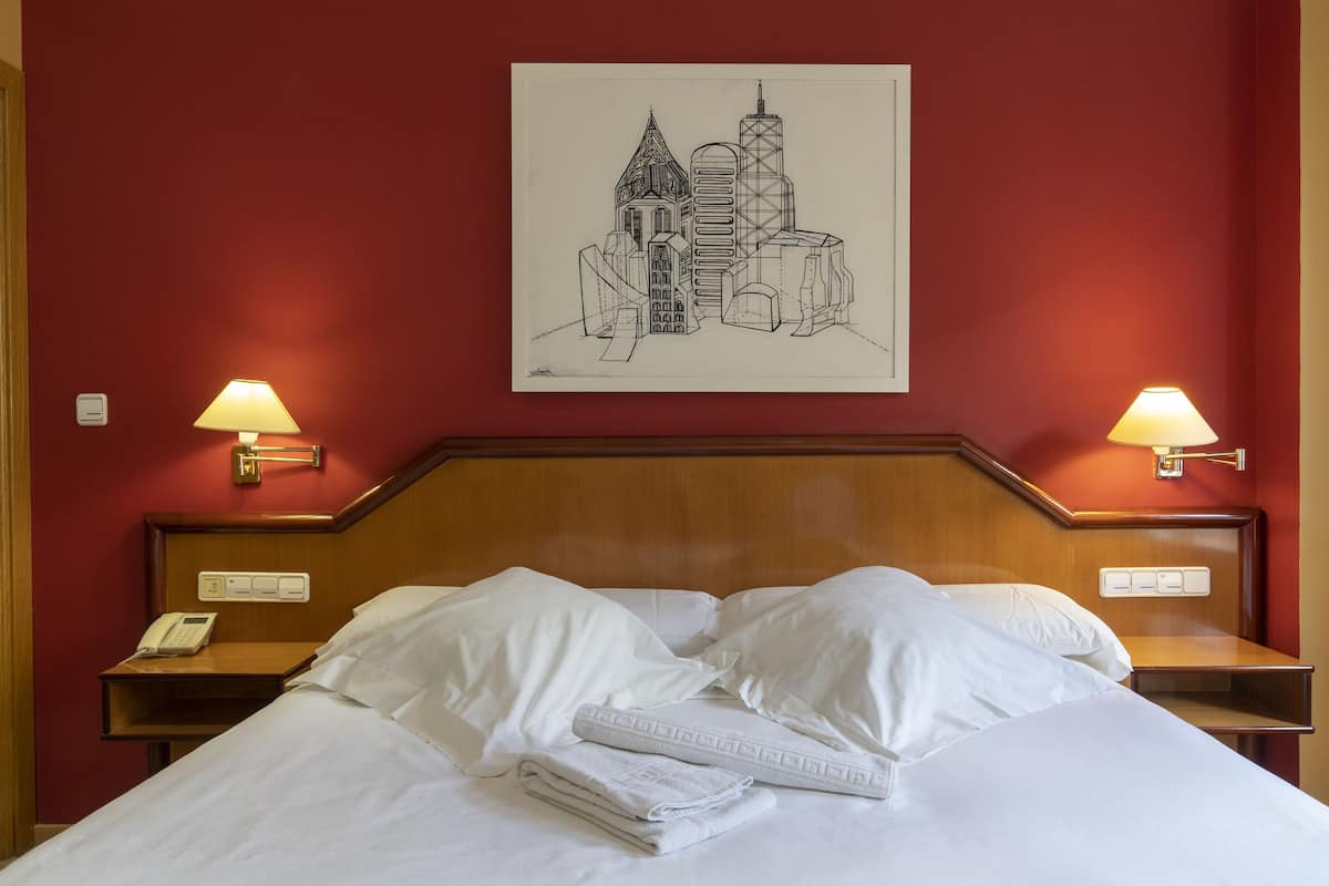 A hotel room with a bed, white linens, a nightstand with a phone, two wall-mounted lamps, and a framed cityscape artwork.
