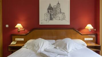 Hotellihuone, jossa on sänky, valkoiset vuodevaatteet, yöpöytä, jossa on puhelin, kaksi seinään kiinnitettyä lamppua ja kehystetty kaupunkimaisemaa esittävä taideteos.