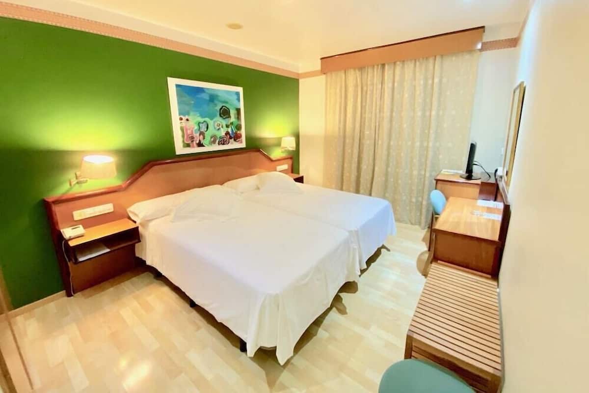 A hotel room with a large bed, two bedside tables, a desk with a chair, a painting on the wall, and a window with curtains.