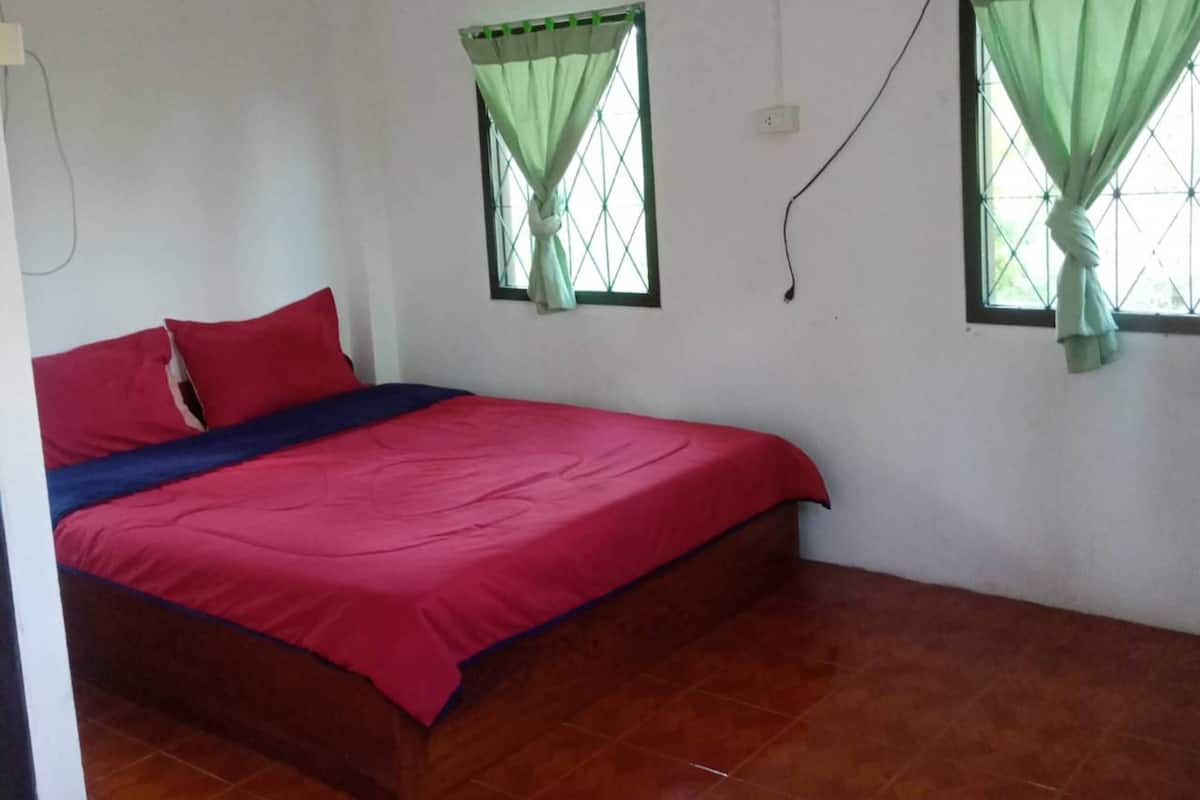 A bedroom with a bed, two windows with curtains, a fan, and a tiled floor.