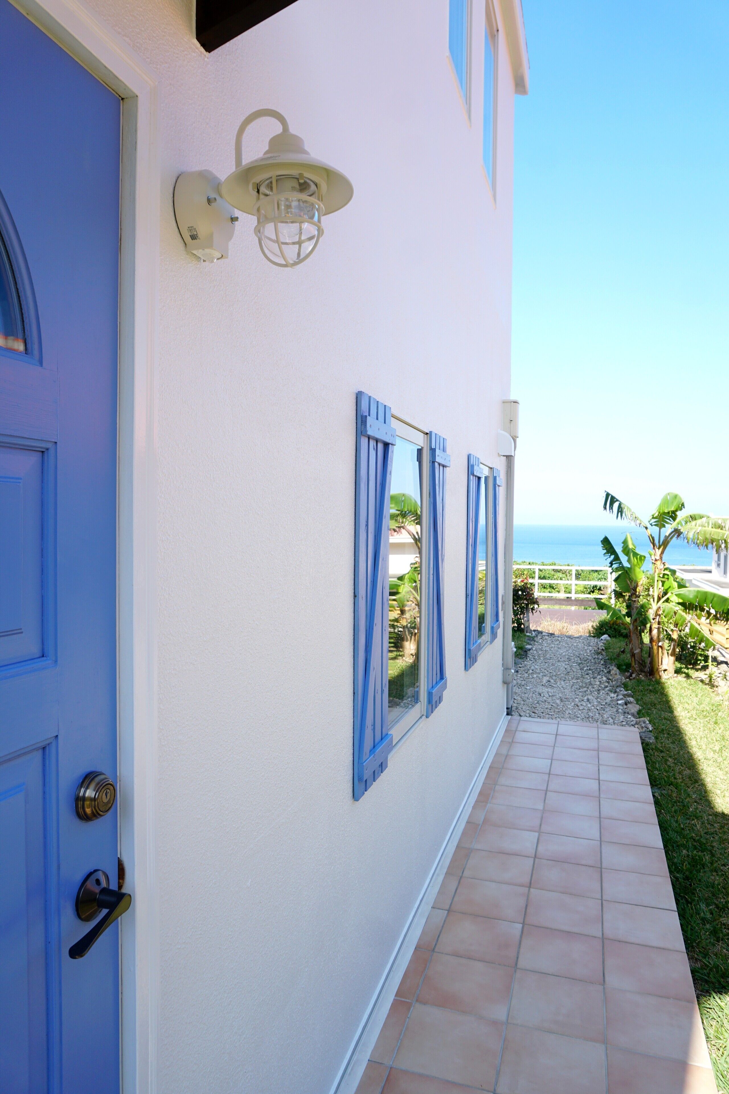 A coastal building with a blue door, white walls, and a tiled pathway leading to a balcony with a view of the sea.