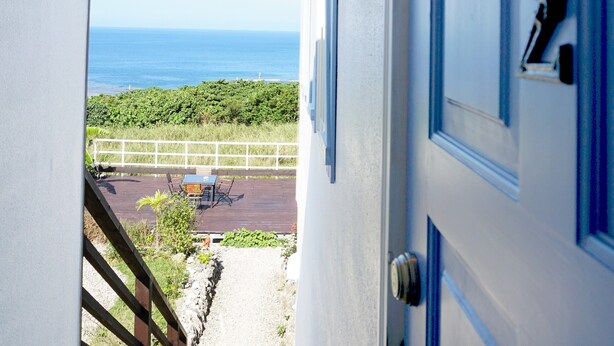 Una puerta azul con una placa de número, vista al mar y un patio con muebles de exterior.