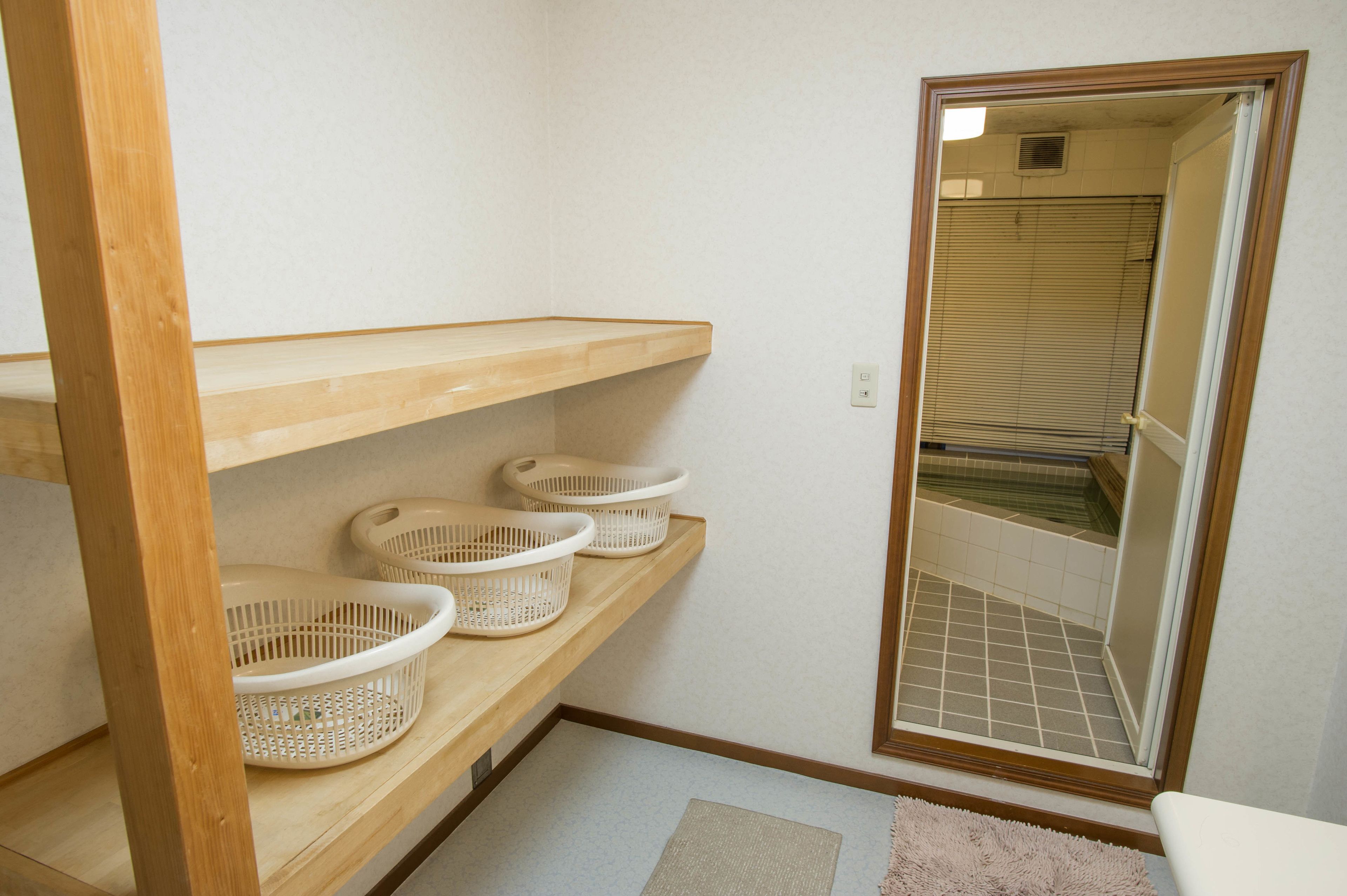 A bathroom with wooden shelves holding white baskets, a mirror reflecting a tiled shower area, and a light-colored rug on the floor.