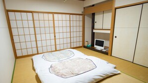 Una habitación japonesa tradicional con suelo de tatami, un futón con almohadas decorativas, un biombio shoji, un escritorio pequeño con una computadora y un armario.