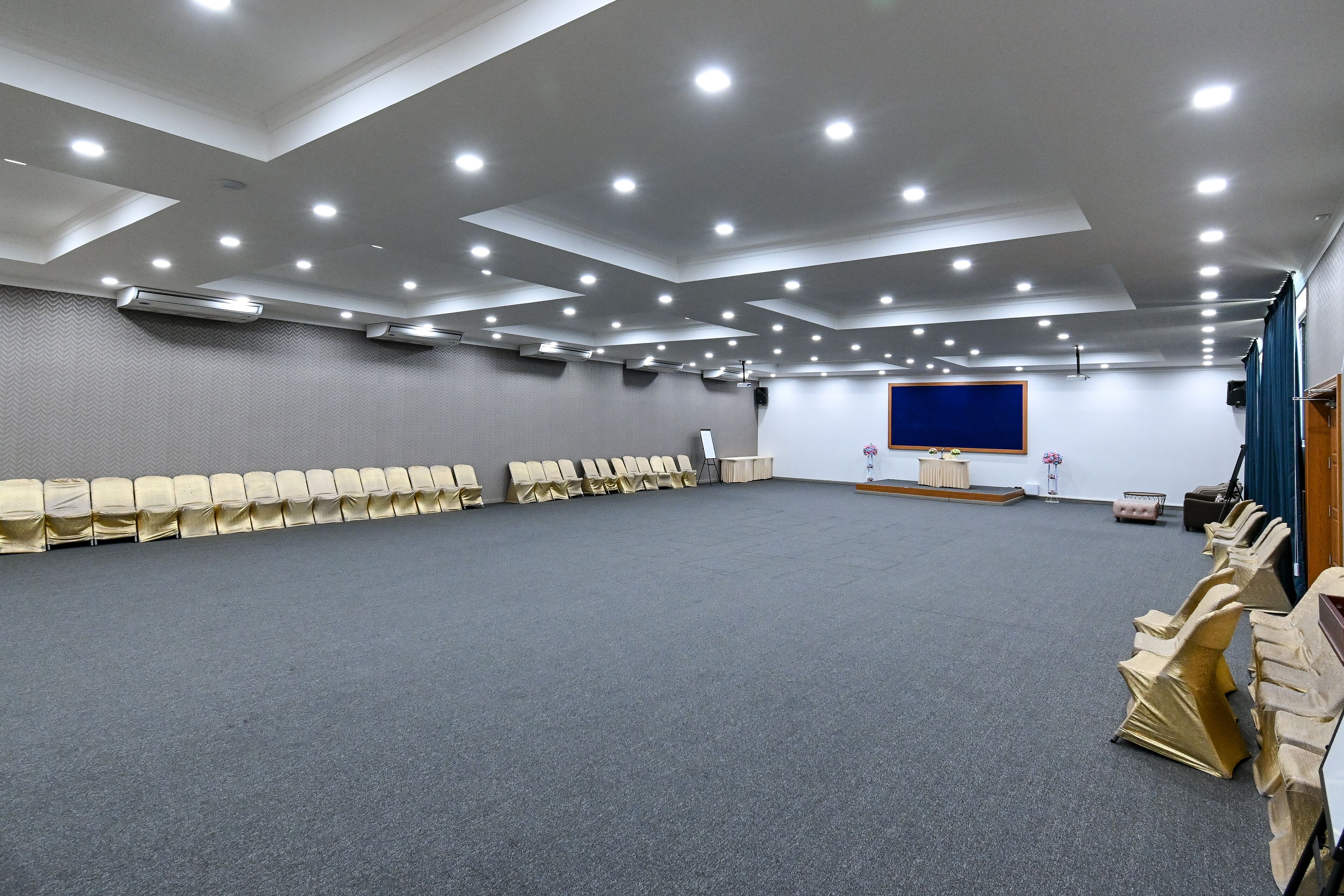Une grande salle de conférence, bien éclairée, avec des rangées de chaises beiges, une scène dotée d’un fond bleu et un sol recouvert d’un tapis gris.