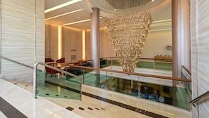 Een moderne hotel lobby met een glazen trap, een grote kroonluchter en een zithoek met rode stoelen.