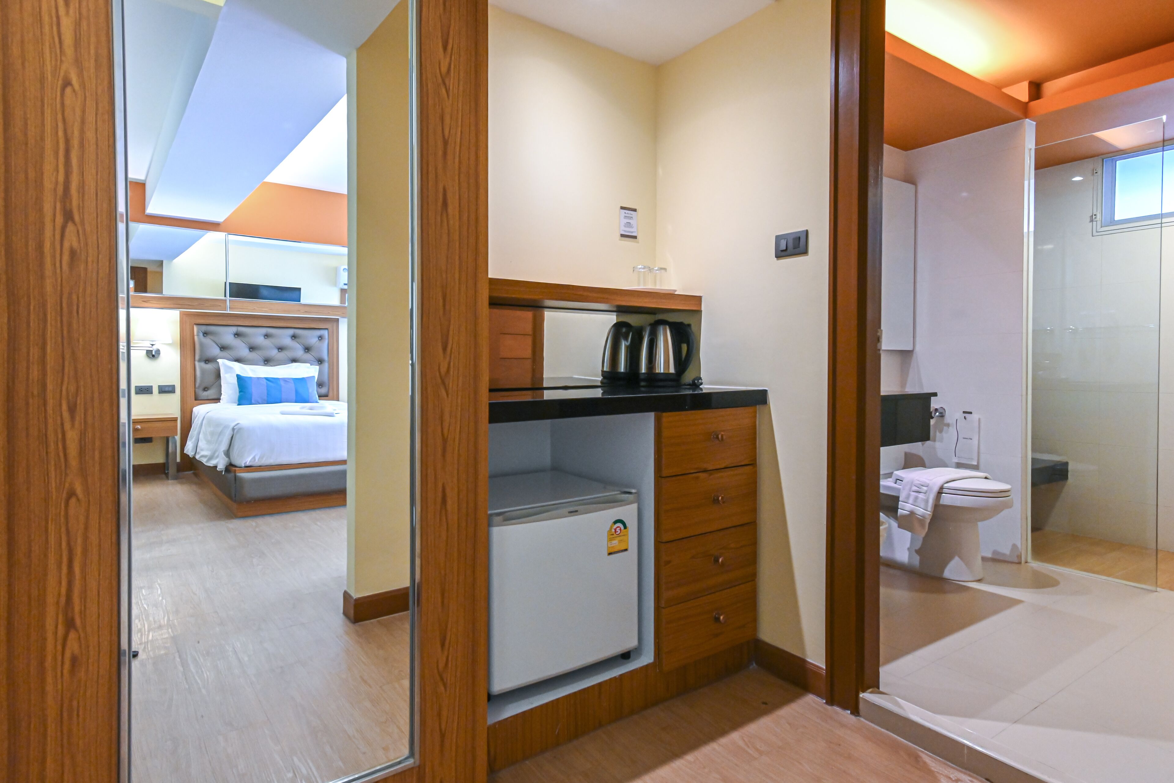 A compact hotel room with a bed, a small kitchenette, a bathroom, and a glass shower.