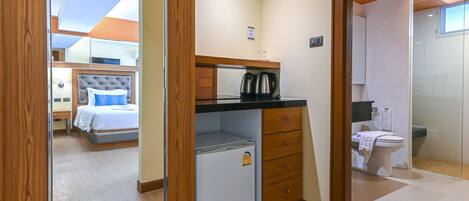A compact hotel room with a bed, a small kitchenette, a bathroom, and a glass shower.