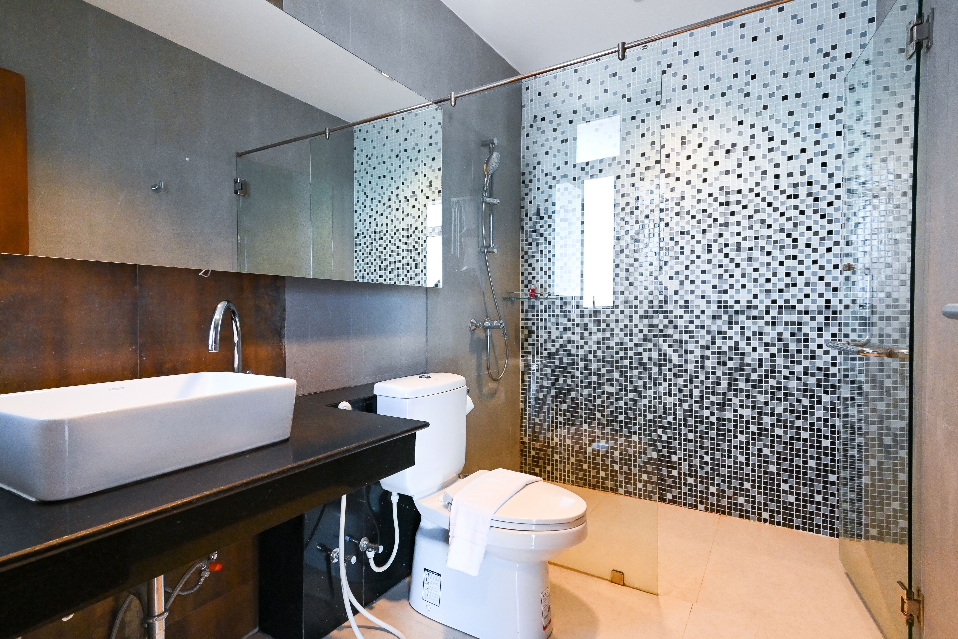 A modern bathroom with a large mirror, a white sink, a toilet, and a shower area with mosaic tiles.