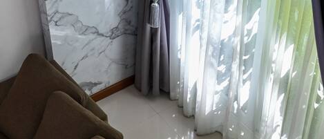 Una habitación con pared de mármol, sofá marrón y cortinas traslúcidas que dejan entrar la luz natural.
