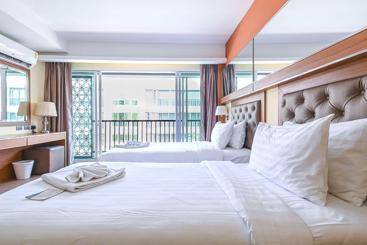 A hotel room with a large bed, a bedside table with a lamp, a window with a view of buildings, and a balcony with a railing.