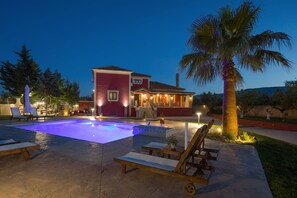 A pool area with lounge chairs, a palm tree, and a building with a red facade. - Villa Froso (Zakynthos)