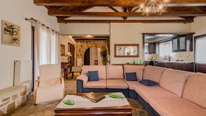 A cozy living room with a stone fireplace, a large beige sofa, a wooden coffee table, and a dining area with a chandelier. - Villa Froso (Zakynthos)