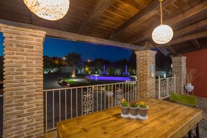 An outdoor patio area with a wooden table, potted plants, and a view of a lit fountain and pool at night. - Villa Froso (Zakynthos)