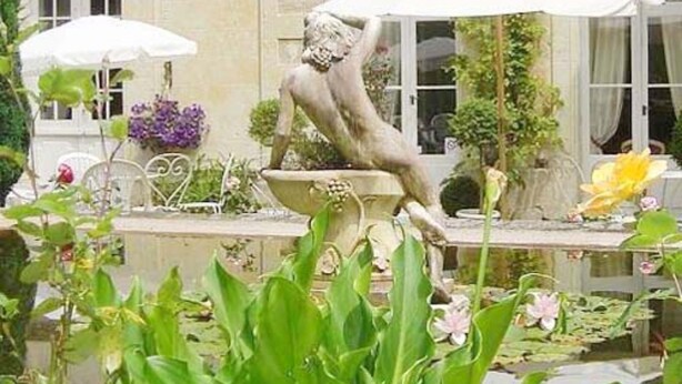 A statue in a garden with a fountain, surrounded by lush greenery and a building with windows in the background. - Château de Beaulieu (Saumur)